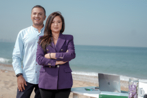 A man and woman stand on the beach, each using a laptop while enjoying the ocean view.