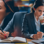 A woman in a business suit drinks coffee while typing on her laptop at a desk.