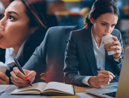 A woman in a business suit drinks coffee while typing on her laptop at a desk.