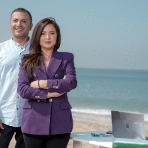 A man and woman stand on the beach, each using a laptop while enjoying the ocean view.